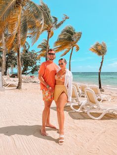 a man and woman standing on the beach in front of palm trees with chairs around them