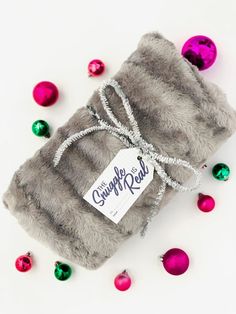 a gray blanket wrapped in silver and pink christmas ornaments on a white surface with a tag