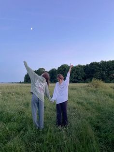 two people standing in a field holding hands and reaching up to the sky with trees behind them