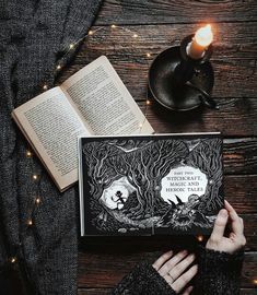 a person holding an open book on top of a wooden table next to a candle