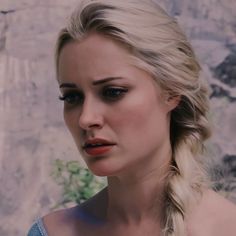 a woman with blonde hair and blue eyes stares into the distance while standing in front of a stone wall