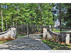 an iron gate with stone pillars leads into a driveway surrounded by trees and shrubs in front of a house