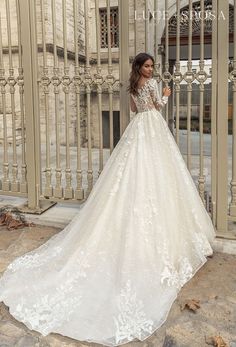 a woman standing in front of a gate wearing a white wedding dress with long sleeves