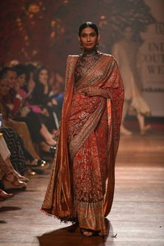 a model walks down the runway in a red and gold sari with an intricate shawl