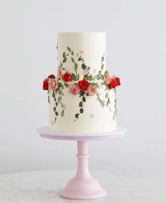 a three tiered cake decorated with flowers and greenery on a pink stand against a gray background