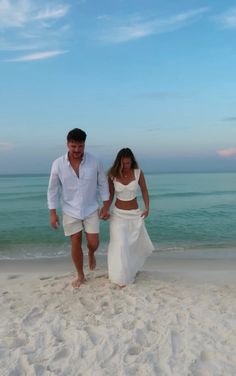 a man and woman are walking on the beach holding hands while dressed in white clothes