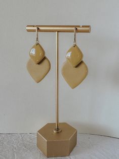 a pair of beige heart shaped earrings are hanging from a gold metal stand on a white surface