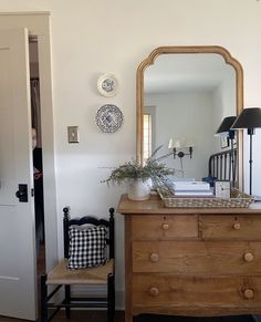 a room with a dresser, mirror and lamp on it's side table in front of a door