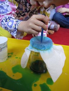 a young child is painting with blue and green paint on a yellow tablecloth while another child sits in the background