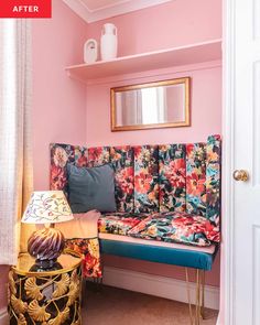 a pink room with a colorful floral couch and mirror on the wall, next to a yellow table lamp