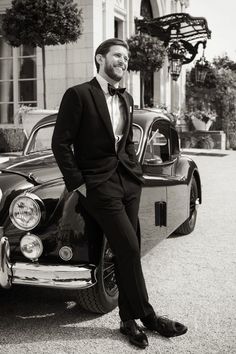 a man in a tuxedo leaning on the hood of an old classic car