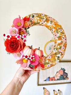 a person holding up a wreath made out of paper flowers and pumpkins in front of a mirror