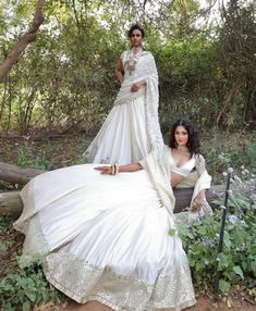 two women in white dresses sitting on a bench with trees and bushes behind them, posing for the camera