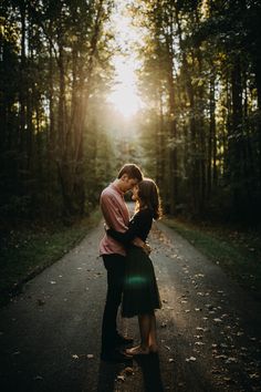 a man and woman standing in the middle of a road surrounded by trees with the sun shining down on them