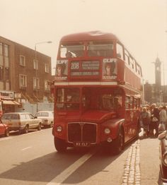 a red double decker bus driving down a street next to parked cars and people walking on the sidewalk