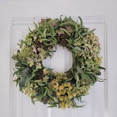 a wreath hanging on the front door of a house with green leaves and flowers around it