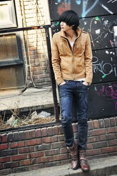 a man standing in front of a brick wall wearing a brown jacket and blue jeans