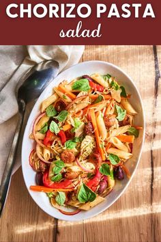 a white bowl filled with pasta and vegetables on top of a wooden table next to a spoon