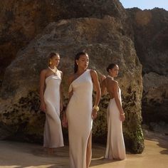 three women in white dresses standing on the beach next to large rocks and boulders, with one wearing a gold bracelet