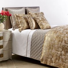 a large bed with gold and white comforters on it's sides, next to a nightstand with a flower in a vase