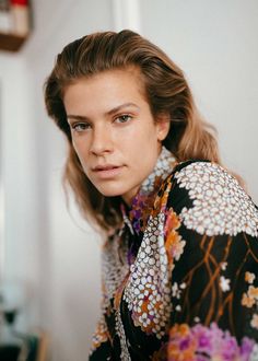 a woman with long hair wearing a floral shirt and looking at the camera while standing in front of a white wall