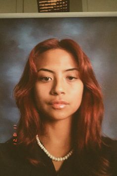 a woman with red hair and pearl necklace in front of a television screen showing an image of her face