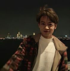 a young man standing next to the ocean at night with city lights in the background