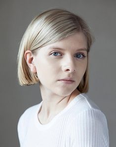 a woman with blonde hair wearing a white shirt and looking at the camera while standing in front of a gray background