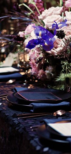 the table is set with purple flowers and black placemats, silverware, and napkins