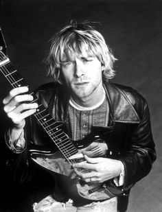 a man holding an electric guitar in his right hand and looking at the camera while wearing a leather jacket