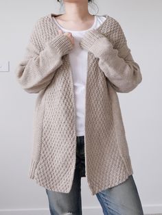 a woman wearing a cardigan sweater and jeans standing in front of a white wall