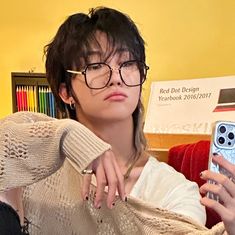 a woman wearing glasses is holding a cell phone and looking at the camera while sitting in front of a bookshelf