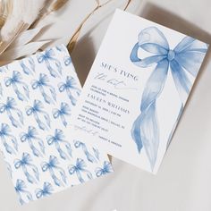 two wedding cards with blue bows on them next to some feathers and flowers in the background