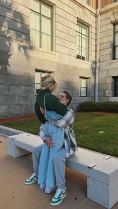 two people hugging each other on a bench in front of a building with grass and trees