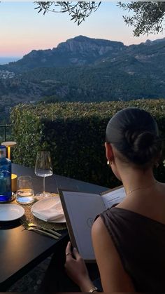 a woman sitting at a table with wine glasses on it looking out over the mountains