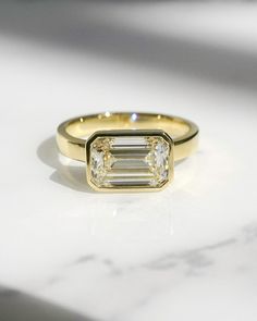 a yellow gold ring with an emerald cut diamond in the center on a marble surface