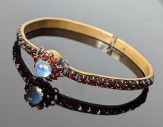 A Victorian Ceylon Blue Moonstone and Garnet Paste Bangle....this piece is set with an extremely blue Ceylon Blue Moonstone as the centre of the flower, surrounded by Rose Cut paste stones.  The bangle has channel set paste garnets running right around the circumference. These stones are deep red but with a brighter red glow.  This piece fastens with a box clasp which is part of the flower- this works well.  It measures 7 cm in diameter and 4.5 mm in width. The central flower is 1.4 cm across an Wedding Bangle Bracelet With Cabochon, Wedding Bracelets With Gemstone Accents, Blue Moonstone, Box Clasp, Channel Set, Etsy Jewelry, Style Gift, Deep Red, Rose Cut