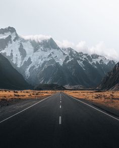 an empty road in the middle of nowhere with snow capped mountains behind it and brown grass on both sides