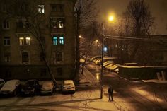 a person walking down a snow covered street at night with cars parked on the side