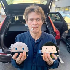 a man standing in front of a car holding up two paper dolls with faces on them