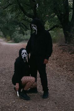 two people dressed in black and white are standing on a dirt road with trees behind them