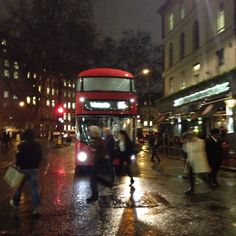 a red double decker bus driving down a city street at night with people crossing the street