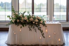 white roses and greenery are arranged on a table with candles in front of large windows