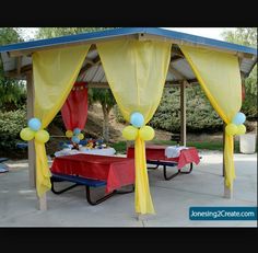 an outdoor picnic area with yellow drapes and red table cloths, blue tables and balloons