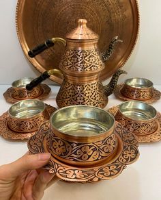 a hand is holding an ornate tea set in front of a plate with four cups and saucers