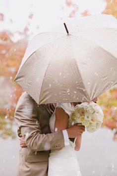 a bride and groom kissing under an umbrella in the rain with fall foliage behind them