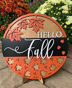 an orange and black sign that says hello fall with leaves on the bottom, in front of some flowers