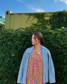 a woman standing in front of bushes wearing a blue jean jacket and pink floral dress