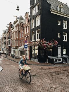 two people riding a bike down a cobblestone street in front of tall buildings