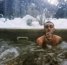 a man in a hot tub drinking from a bottle with snow on the ground behind him